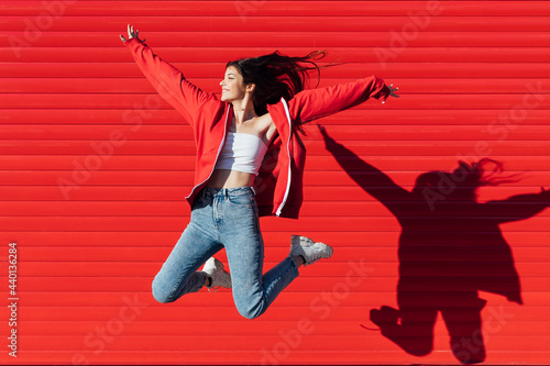Smiling teenage girl with arms outstretched jumping in front of red wall photo
