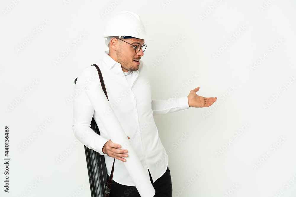 Young architect man with helmet and holding blueprints isolated on white background with surprise expression while looking side Young architect man with helmet and holding blueprints isolated on white background with surprise expression while looking side