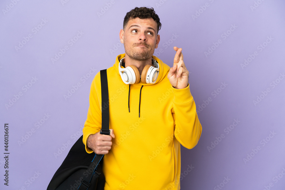 Young sport caucasian man with bag isolated purple background with fingers crossing and wishing the best Young sport caucasian man with bag isolated purple background with fingers crossing and wishing the best