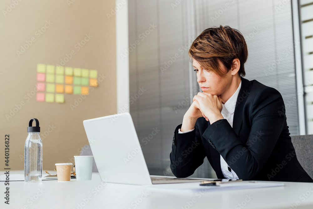 Thoughtful female entrepreneur with hand on chin looking at laptop while sitting in office Thoughtful female entrepreneur with hand on chin looking at laptop while sitting in office