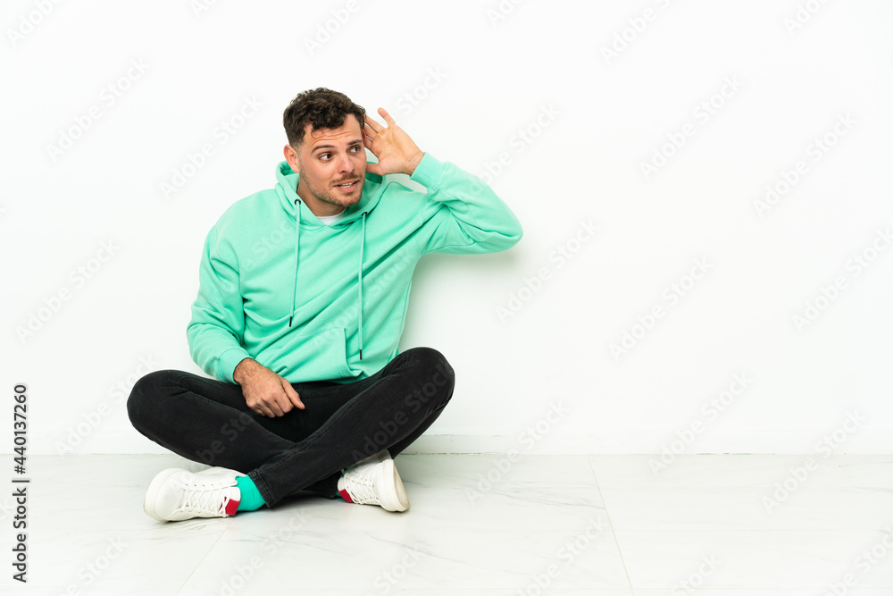 Young handsome caucasian man sitting on the floor listening to something by putting hand on the ear Young handsome caucasian man sitting on the floor listening to something by putting hand on the ear