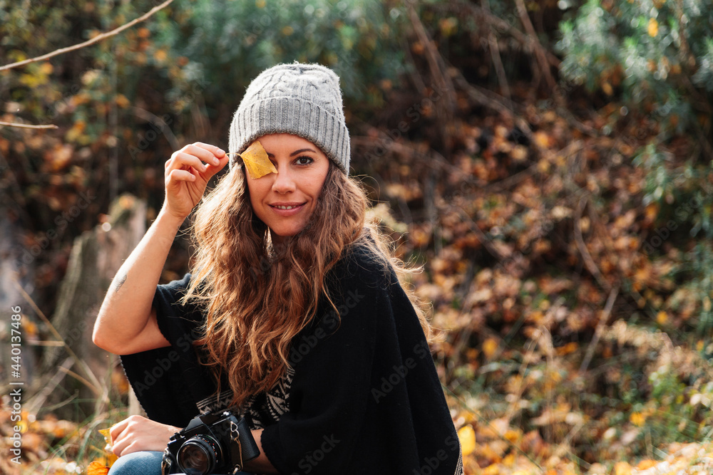 Smiling woman covering eye with autumn leaf while sitting in forest during vacations