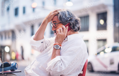 Senior man talking on mobile phone with hand in hair while sitting at sidewalk cafe