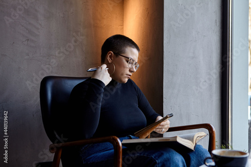 A young girl with glasses, with short hair is sitting in a cafe and reading a book. The girl looks at her book and makes notes in a notebook. The concept of distance learning