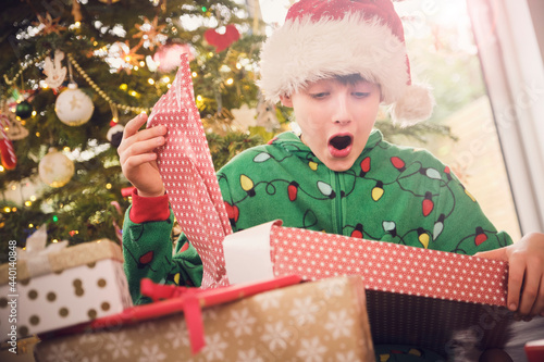 Excited boy in Santa hat unwrapping gift with Christmas tree in background at home