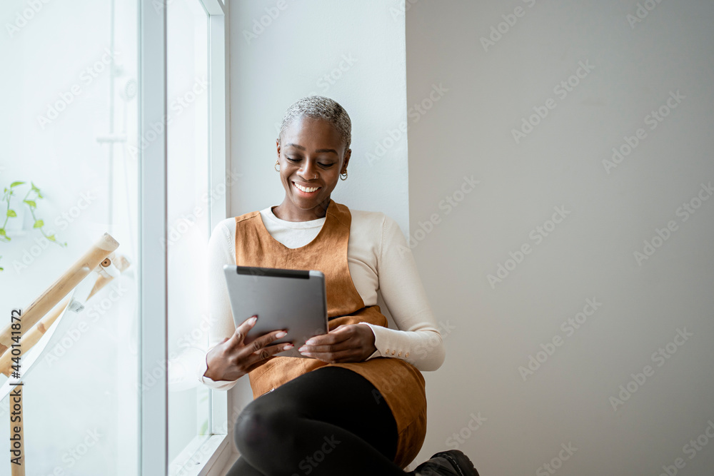 © Rafa Cortes/Westend61 - Smiling woman looking at digital tablet while sitting by window in living room © Rafa Cortes/Westend61 - Smiling woman looking at digital tablet while sitting by window in living room