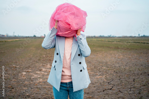 Woman in casuals covering face with pink tulle net on dirt field photo