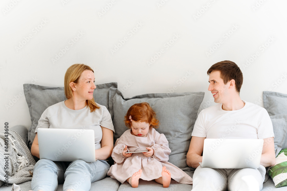 Smiling male and female entrepreneurs with laptop looking at each other while sitting by daughter on sofa in living room Smiling male and female entrepreneurs with laptop looking at each other while sitting by daughter on sofa in living room