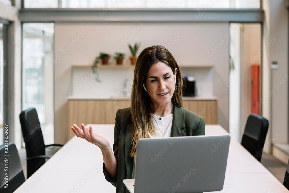 Businesswoman with in-ear headphones gesturing during video call on laptop at office Businesswoman with in-ear headphones gesturing during video call on laptop at office