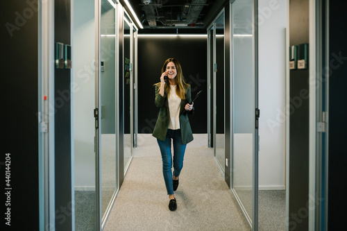 Confident businesswoman talking on smart phone while walking in corridor at office photo