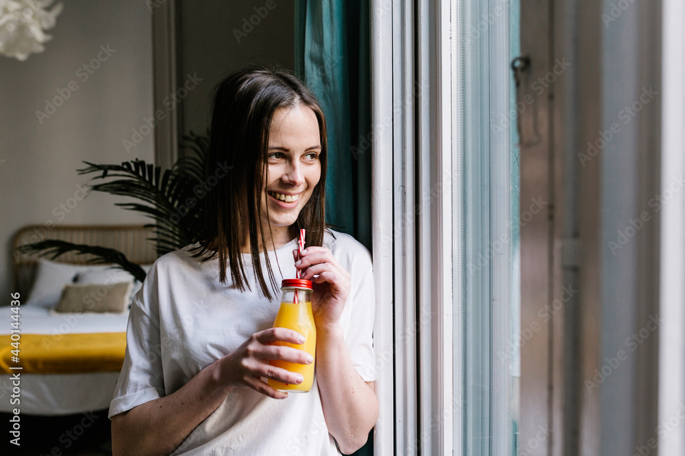 Smiling young woman looking away while having orange juice by window at home Smiling young woman looking away while having orange juice by window at home