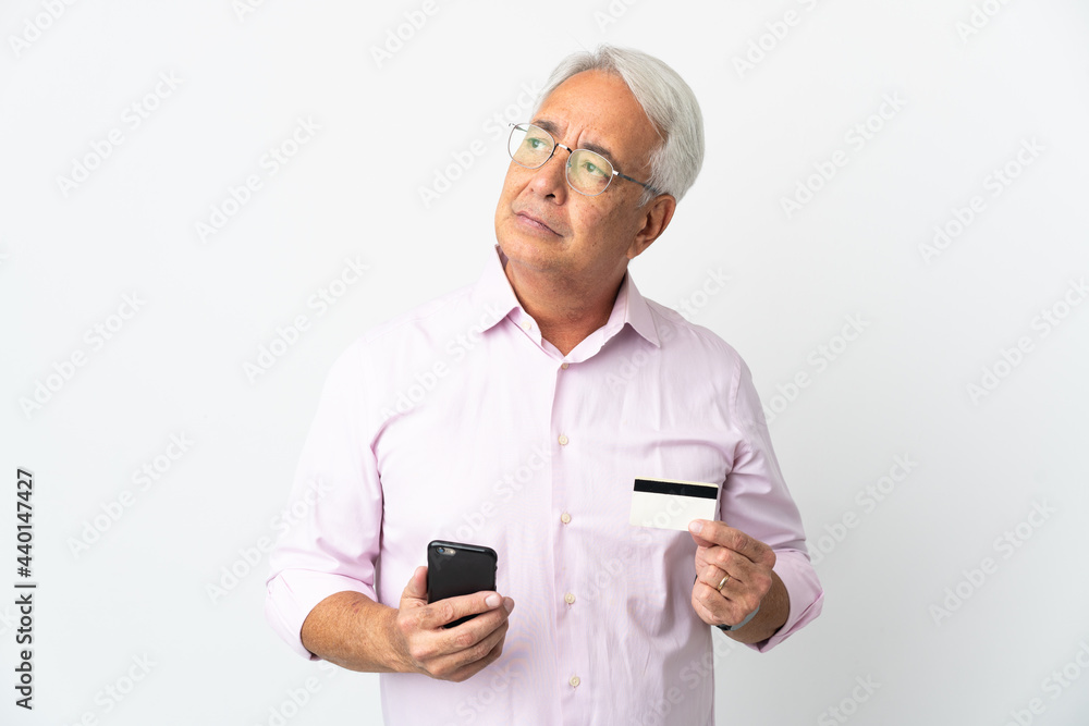 Middle age Brazilian man isolated on white background buying with the mobile with a credit card while thinking Middle age Brazilian man isolated on white background buying with the mobile with a credit card while thinking