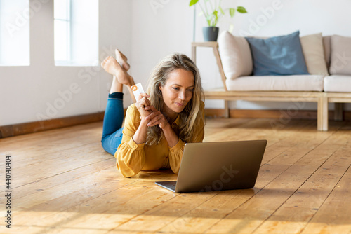 Blond woman with credit card lying down on floor while using laptop at home photo