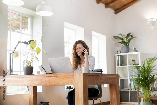 Smiling businesswoman talking on mobile phone while sitting in front of laptop at home office photo