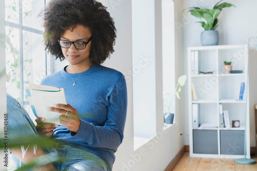 Young woman reading book while sitting by window at home