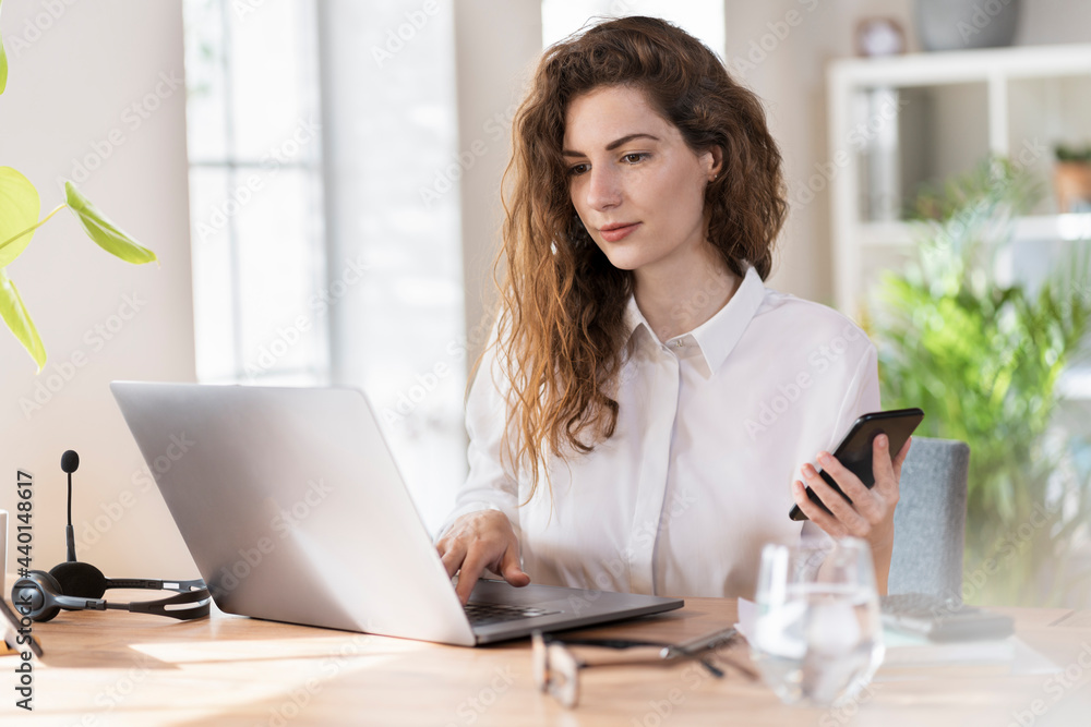 Female professional with mobile phone working on laptop at desk Female professional with mobile phone working on laptop at desk