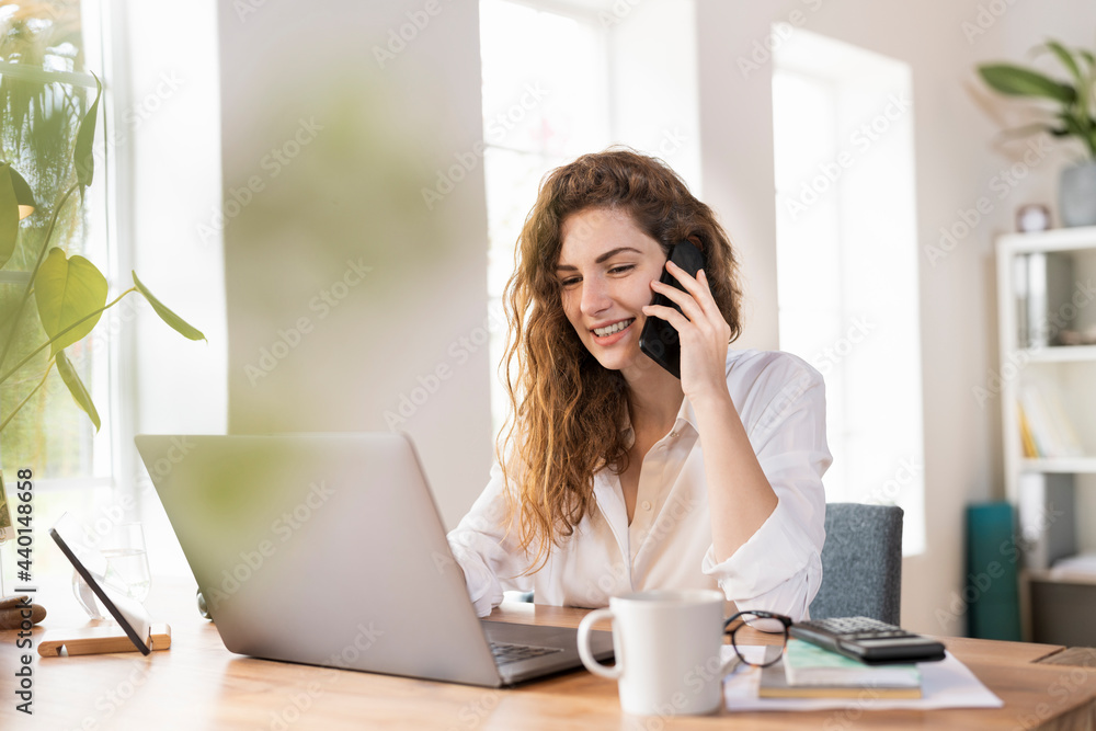 Contented female entrepreneur talking on smart phone while using laptop at desk in home office Contented female entrepreneur talking on smart phone while using laptop at desk in home office