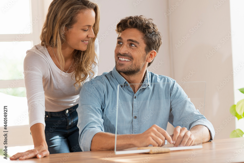 Happy couple looking at each other sitting with transparent screen at home Happy couple looking at each other sitting with transparent screen at home