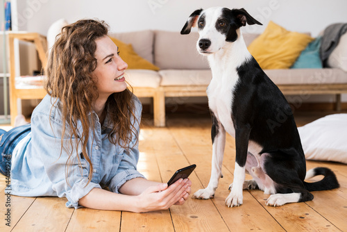 Beautiful woman with long hair holding smart phone while looking at dog in living room photo
