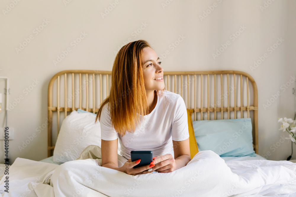 Relaxed woman looking away while holding mobile phone on bed at home Relaxed woman looking away while holding mobile phone on bed at home