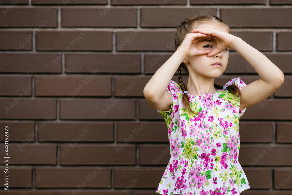 The little girl covered her eyes with her hands and peeks. A cute child in bright clothes stands against a brick wall and discreetly watches. Espionage or covert surveillance concept The little girl covered her eyes with her hands and peeks. A cute child in bright clothes stands against a brick wall and discreetly watches. Espionage or covert surveillance concept