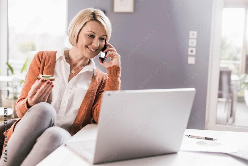 Smiling female entrepreneur talking on smart phone and having food while working at home Smiling female entrepreneur talking on smart phone and having food while working at home