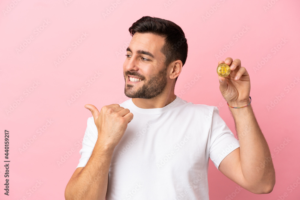 Young man holding a Bitcoin isolated on pink background pointing to the side to present a product Young man holding a Bitcoin isolated on pink background pointing to the side to present a product