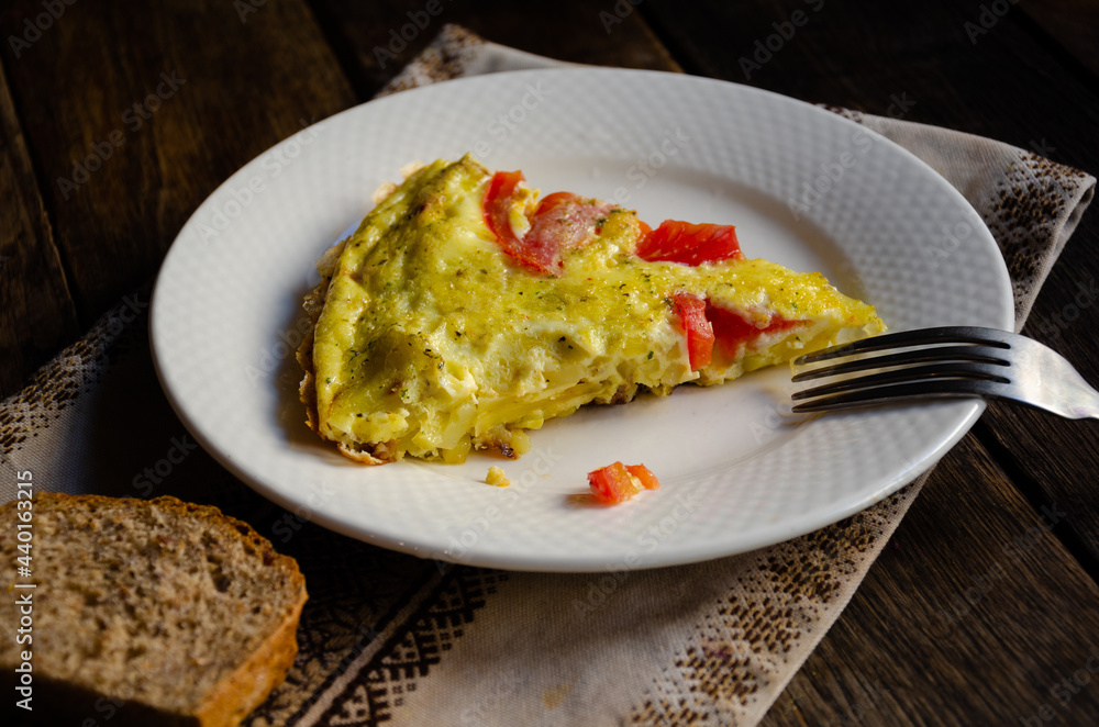 A serving of rustic omelet in a white plate, bread, kitchen napkin on a wooden table.