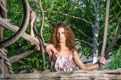 Holding on rattans in woods, a pretty woman with long brown curly hair is confidently looking at you. photo