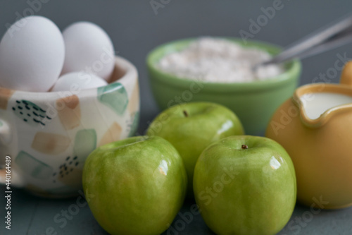Green apples, milk, flour, white eggs in a colorful bowl on a textured gray background
