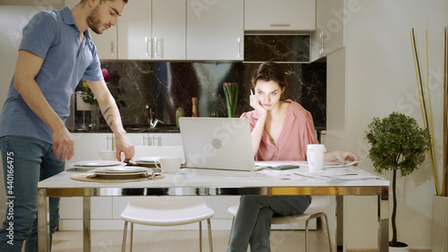An adult woman is sitting and working at home while her husband is doing household chores