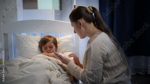 Mother sitting next to sick son in bead and typing doctor phone number on smartphone. Concept of children illness, disease and parent care