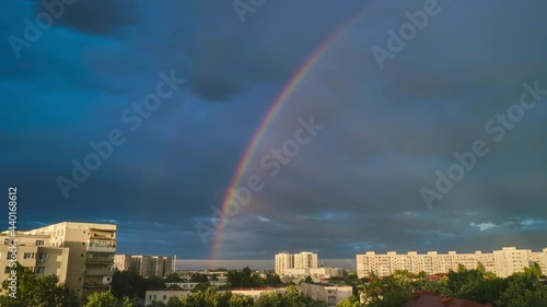 Bright double rainbow and light rays above urban buildings, against dark, blue, after rain sky, in Bucharest, Romania. Timelapse.