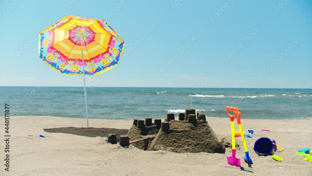 Colourful sunshade next to sandcastle built on beach, plastic children toys around. Sea waves splashing in background. Sunny summer day at seaside. Colourful sunshade next to sandcastle built on beach, plastic children toys around. Sea waves splashing in background. Sunny summer day at seaside.