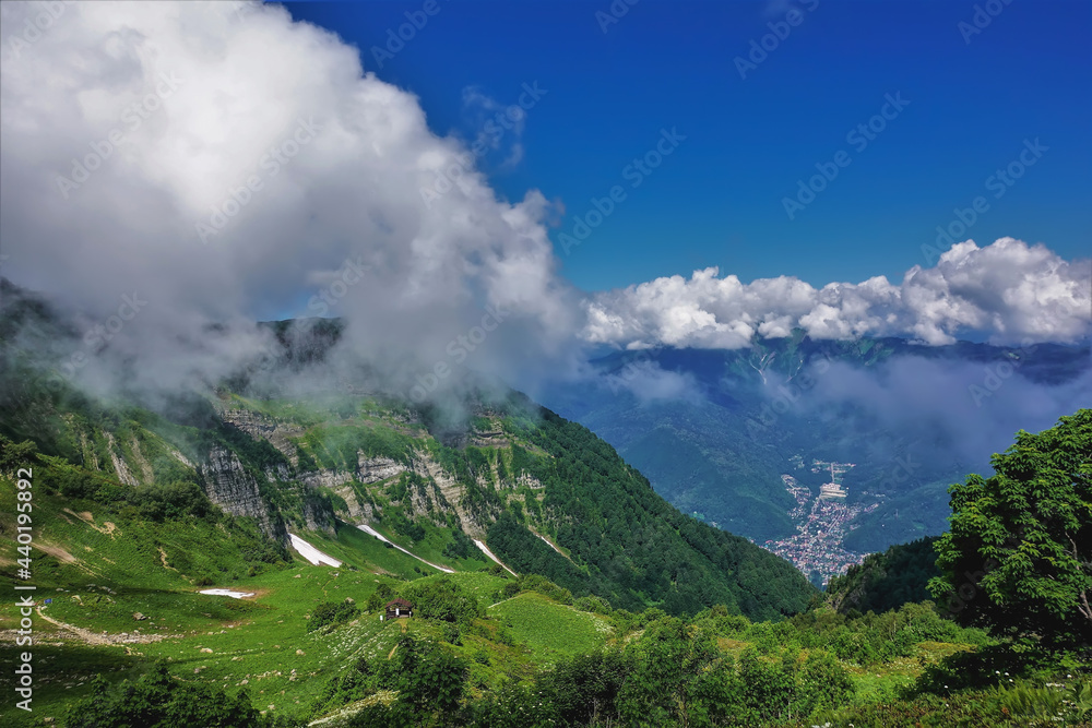 Alpine landscape in summer. In the valley there is green vegetation, areas of snow. The peaks of the mountains are hidden in the clouds. A village in the distance. Caucasus. Russia. Krasnaya Polyana