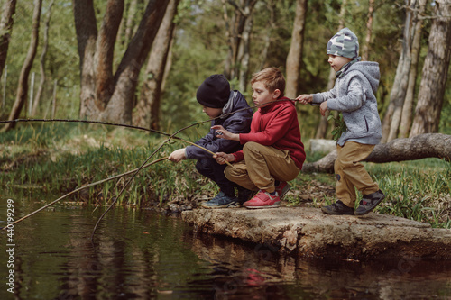 adorable caucasian boys sitting on their hunkers on bank of the pond in forest pretending to do fishing with long branches. Happy childhood concept