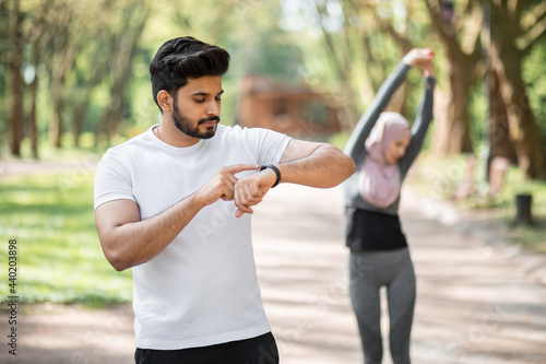 Fototapeta Naklejka Na Ścianę i Meble -  Handsome muslim man using app for training on smart watch while woman in hijab stretching body on background. Young active couple enjoying outdoors workout.