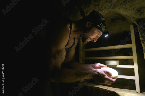 Farmer with headlamp looking at handmade cheese in cellar 