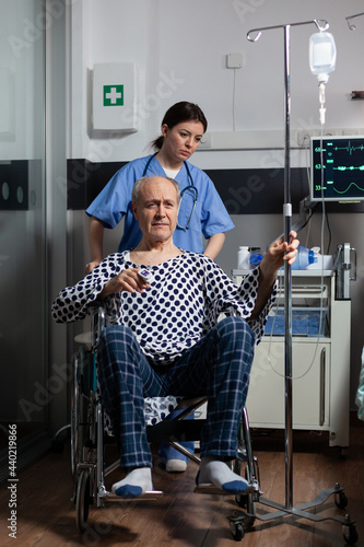 Hospitalized senior man sitting in wheelchair in hospital room, holding iv drip with oximeter attached on finger. Medcal assistant preparing sick patient for treatment. photo