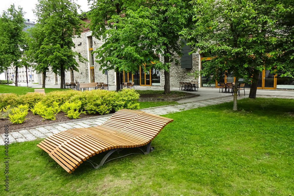 A place to eat and rest outside the office building in Ulemiste. Curved two wood benches in the front and a tables with chairs on the back. Green plants and trees on a summer day. Tallinn, Estonia