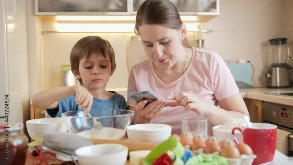 Young mother checking recipe on smartphone while baking pie or cake with her little son. Children cooking with parents, little chef, family having time together, domestic kitchen. Young mother checking recipe on smartphone while baking pie or cake with her little son. Children cooking with parents, little chef, family having time together, domestic kitchen.