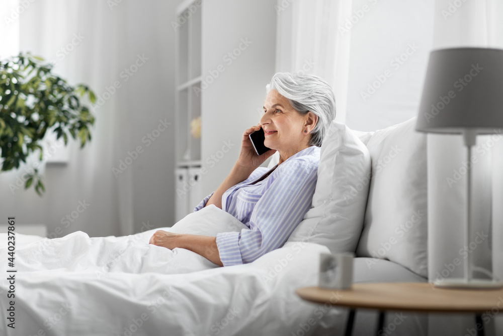 old age, technology and people concept - happy smiling senior woman in pajamas calling on smartphone sitting in bed at home bedroom old age, technology and people concept - happy smiling senior woman in pajamas calling on smartphone sitting in bed at home bedroom