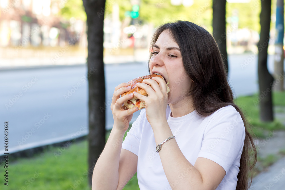 Brunette woman overeats on a burger on the street. Gluttony, excess calories, and bulimia. Brunette woman overeats on a burger on the street. Gluttony, excess calories, and bulimia.