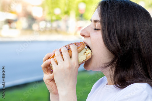 Brunette woman eating burger on the street near the road in the city. Fast food on the way to work or school. Place for text.