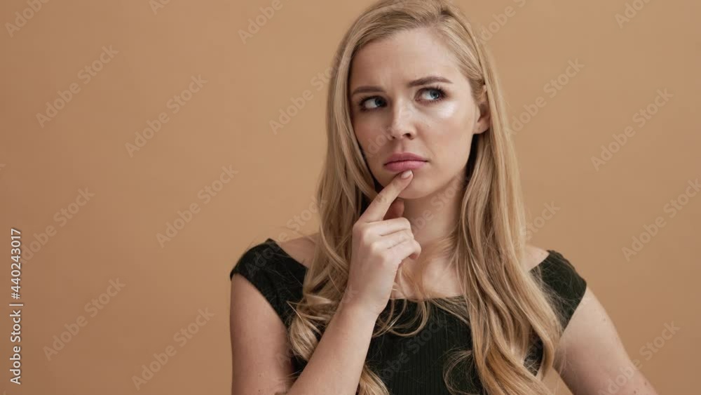 A serious blonde woman is thinking about something standing isolated over a beige wall in the studio A serious blonde woman is thinking about something standing isolated over a beige wall in the studio