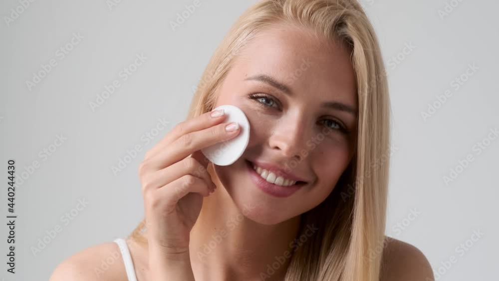 A smiling young blonde woman is wiping her face with a sponge standing isolated over grey wall in the studio A smiling young blonde woman is wiping her face with a sponge standing isolated over grey wall in the studio