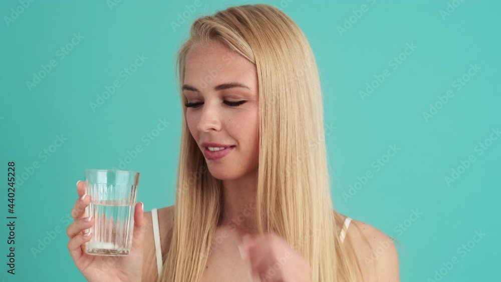 A positive young blonde woman throws a pill into a glass of water standing isolated over blue wall in the studio A positive young blonde woman throws a pill into a glass of water standing isolated over blue wall in the studio