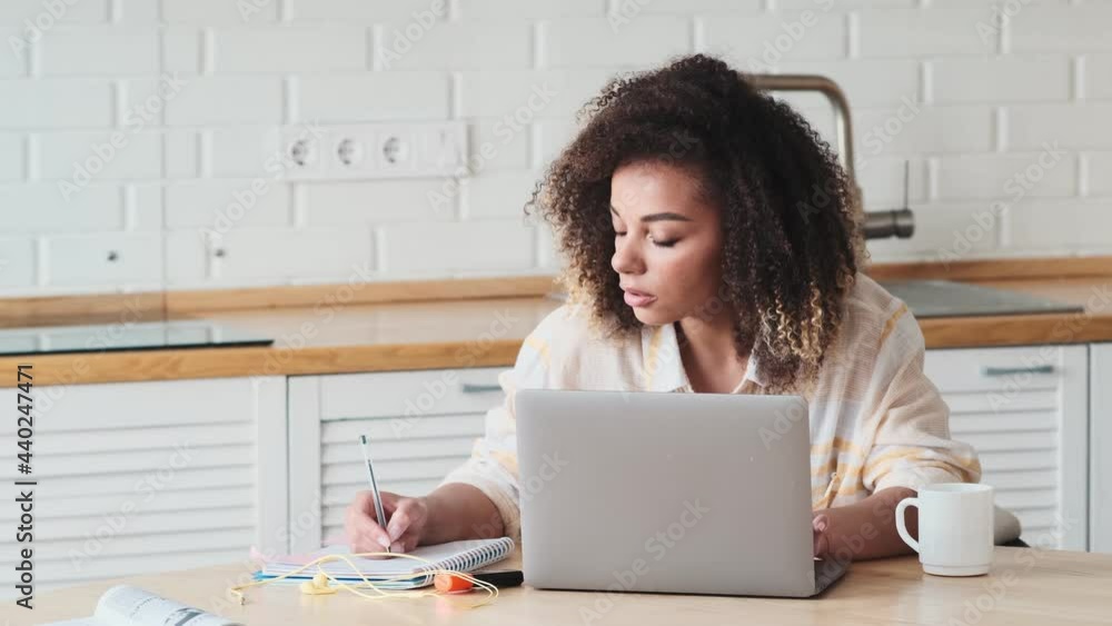 A focused african american woman is writing something while using her laptop computer sitting in the kitchen at home