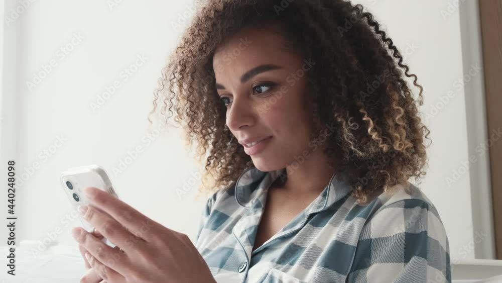 A pleased african american woman wearing pajama is using her smartphone while sitting on the sofa at home A pleased african american woman wearing pajama is using her smartphone while sitting on the sofa at home