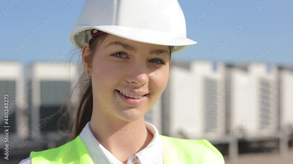 Portrait of a happy woman engineer is looking at the camera. She is smiling. She is wearing work clothes and a hard hat. She's on the roof of a business center. Air conditioning systems in the Portrait of a happy woman engineer is looking at the camera. She is smiling. She is wearing work clothes and a hard hat. She's on the roof of a business center. Air conditioning systems in the